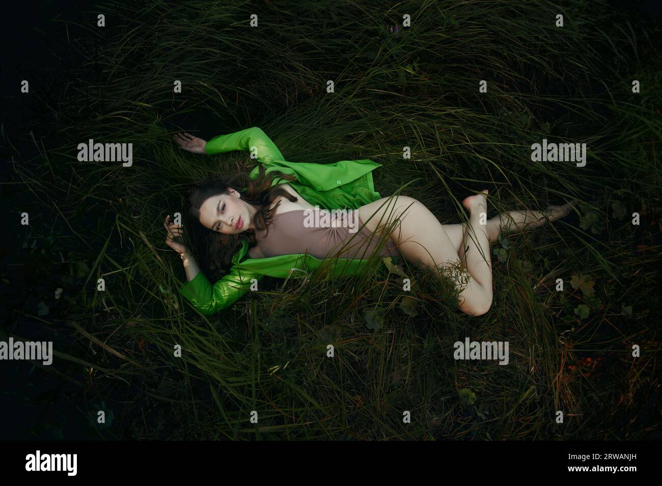 Young woman resting in grass near pond lake. Woman in beige bodysuit and green jacket poses, sensual appearance Stock Photo