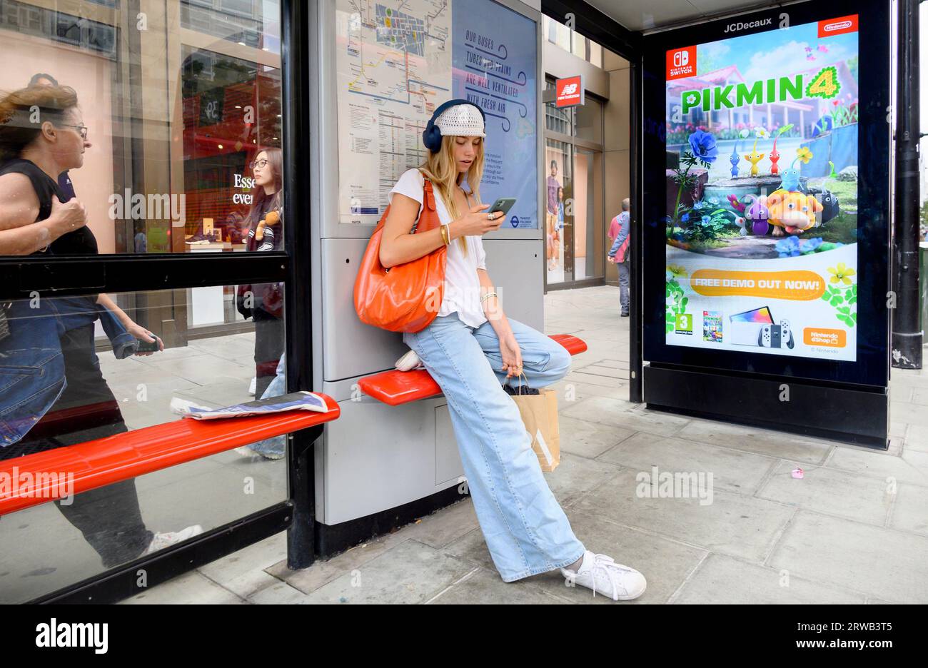 London, UK. Young woman at a bus stop wearing headphones and looking at her phone Stock Photo