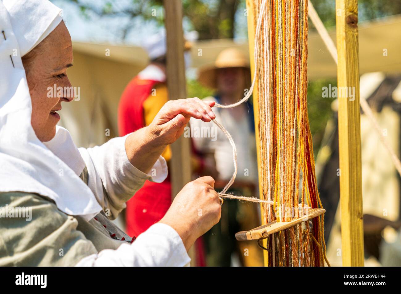 Medieval living history event. Close up of a middle aged lady in middle ages period costume, working on a loom weaving threads of cloth. Stock Photo
