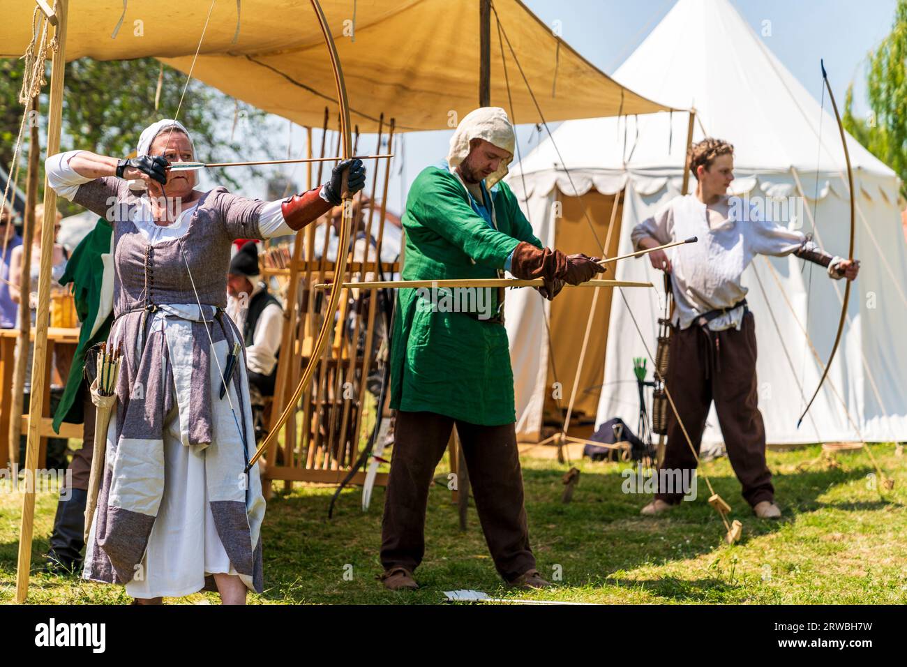Medieval reenactment with a woman in period costume, aiming a bow and arrow, while two men behind her ready their bows. People watching in background. Stock Photo