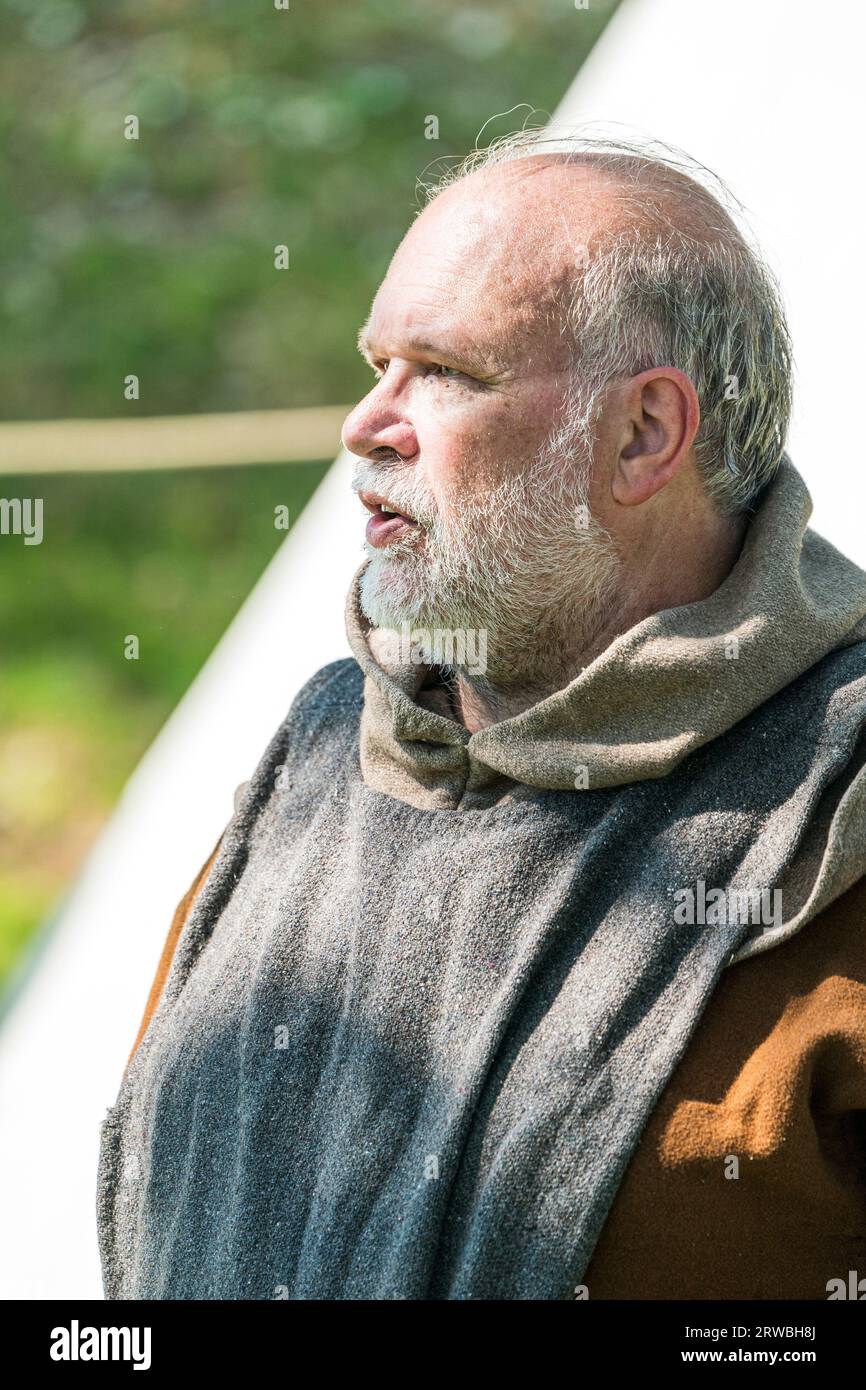 A man dressed as a medieval Friar, monk, or priest during a re-enactment. Side view of the man,  who has grey and brown robes and a grey beard. Stock Photo