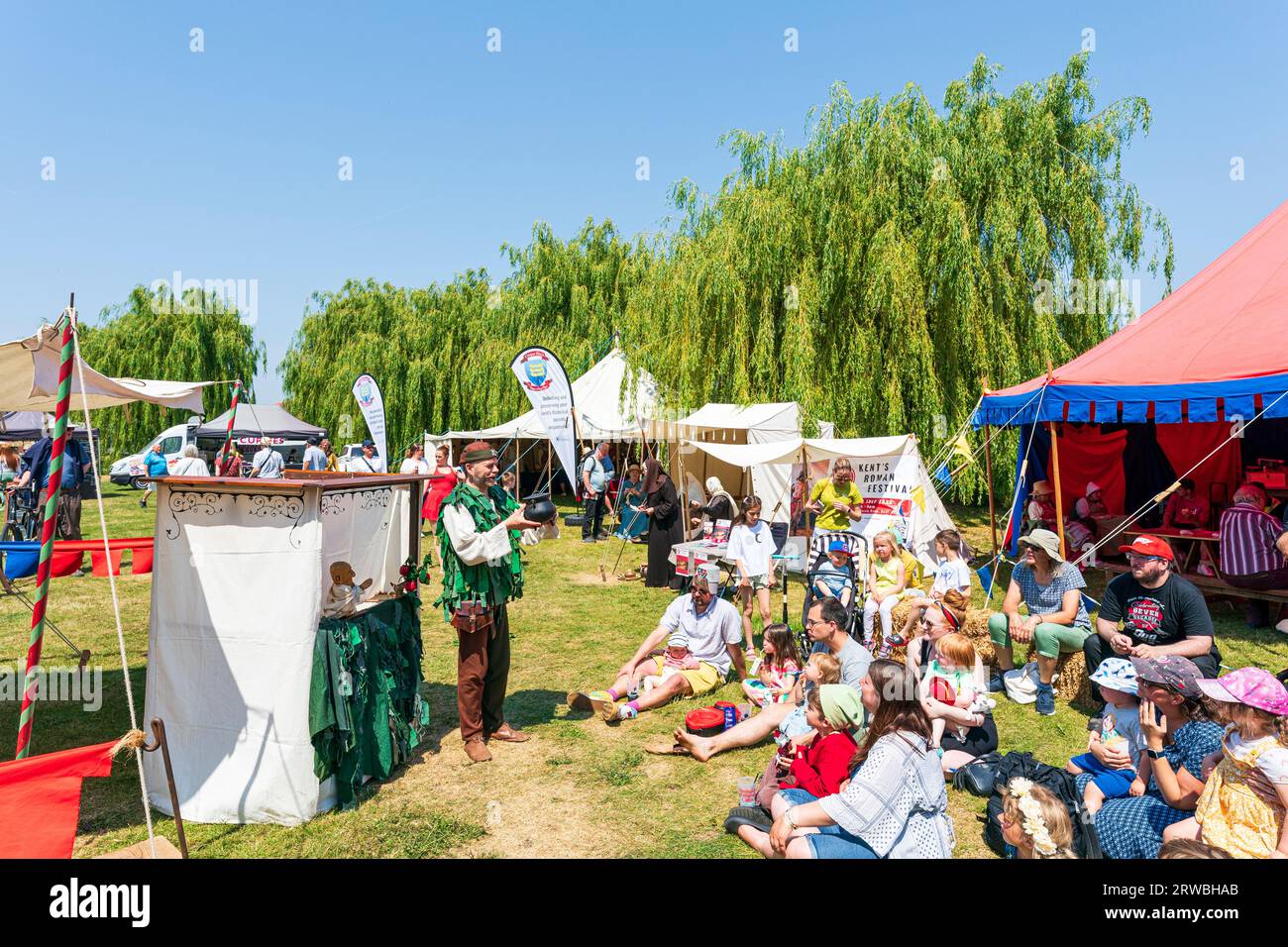 Medieval puppeteer in a green period costume, performing in front of a mainly seated audience on the grass on a hot summer day in bright sunshine. Stock Photo