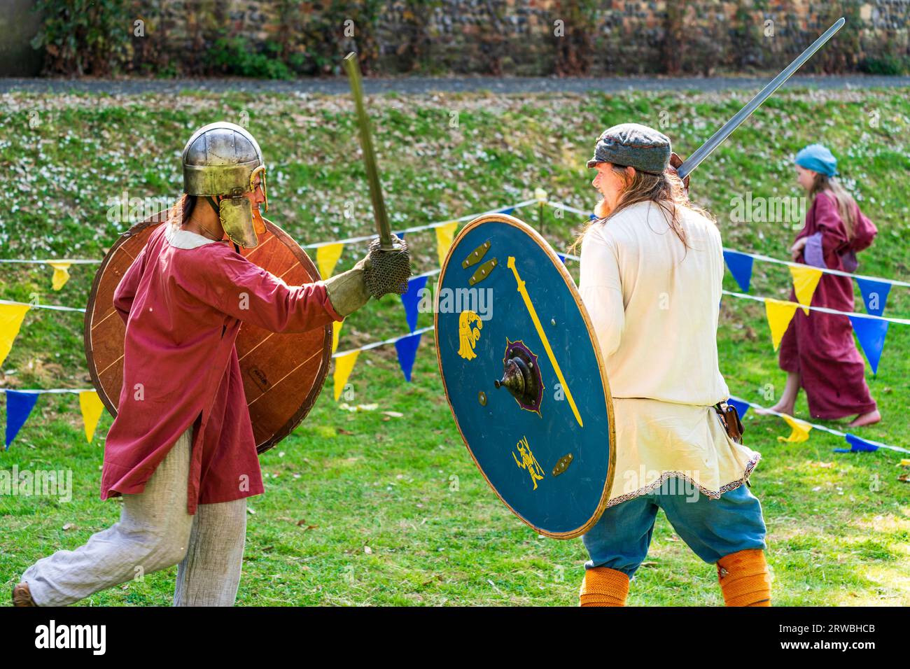 Two men in period costume, jousting with swords in the sunshine during a medieval re-enactment in an area enclosed by bunting of yellow and blue flags Stock Photo