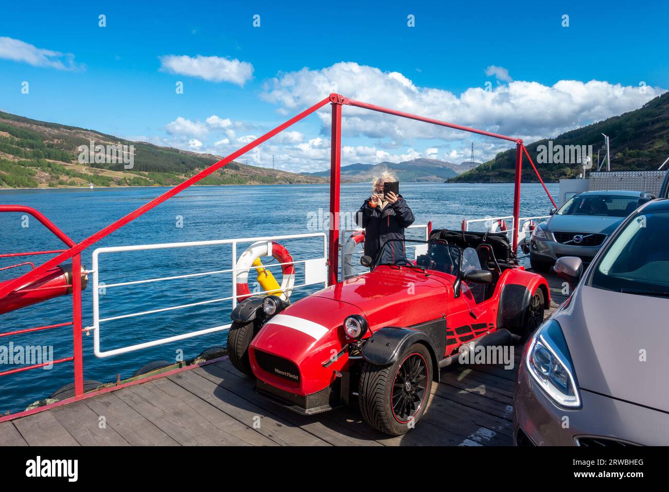 Tourist taking a photo on the Glenelg to Kylerhea Ferry on the Isle of Skye on tour in a red kit car in the Scottish Highlands, Scotland. Stock Photo