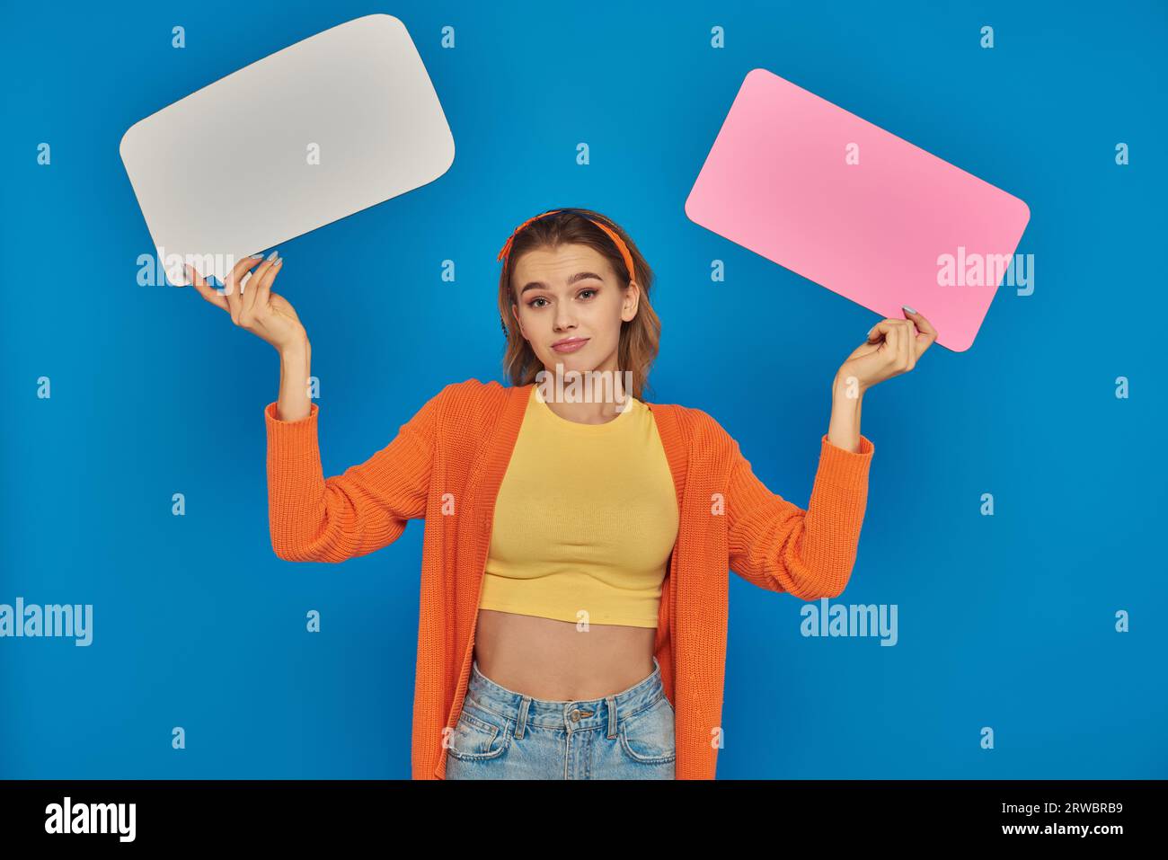 confused young woman in casual attire holding speech bubbles on blue background, space for text Stock Photo