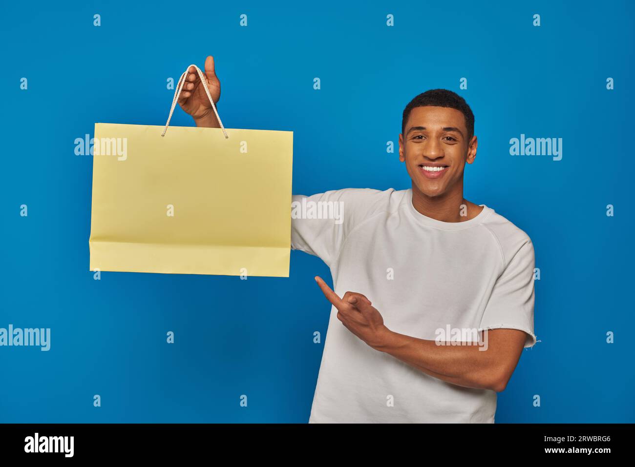 joyful african american man in casual attire pointing at shopping bag on blue backdrop, buying spree Stock Photo
