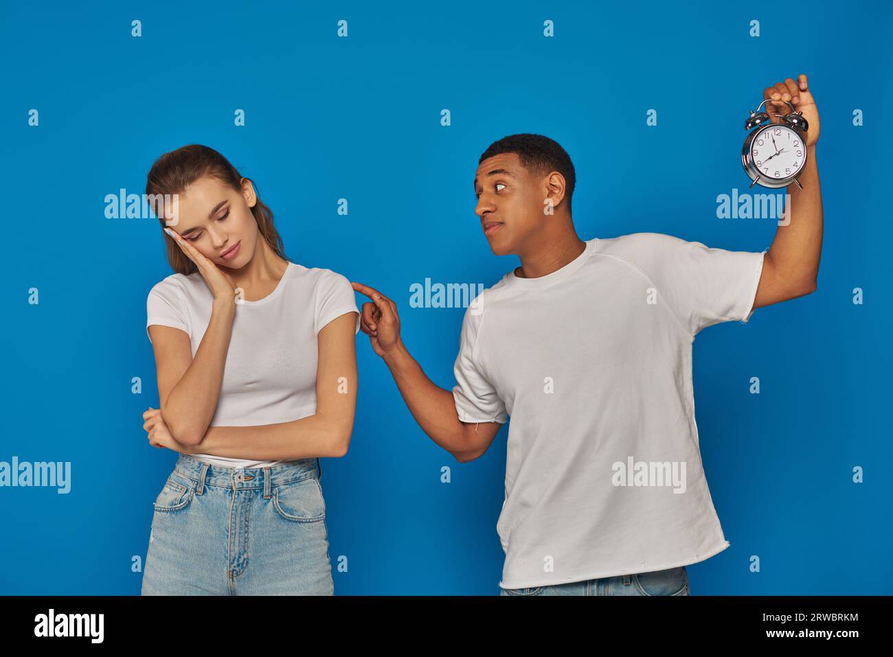 awaken african american man holding alarm clock and waking up sleepy woman on blue backdrop Stock Photo