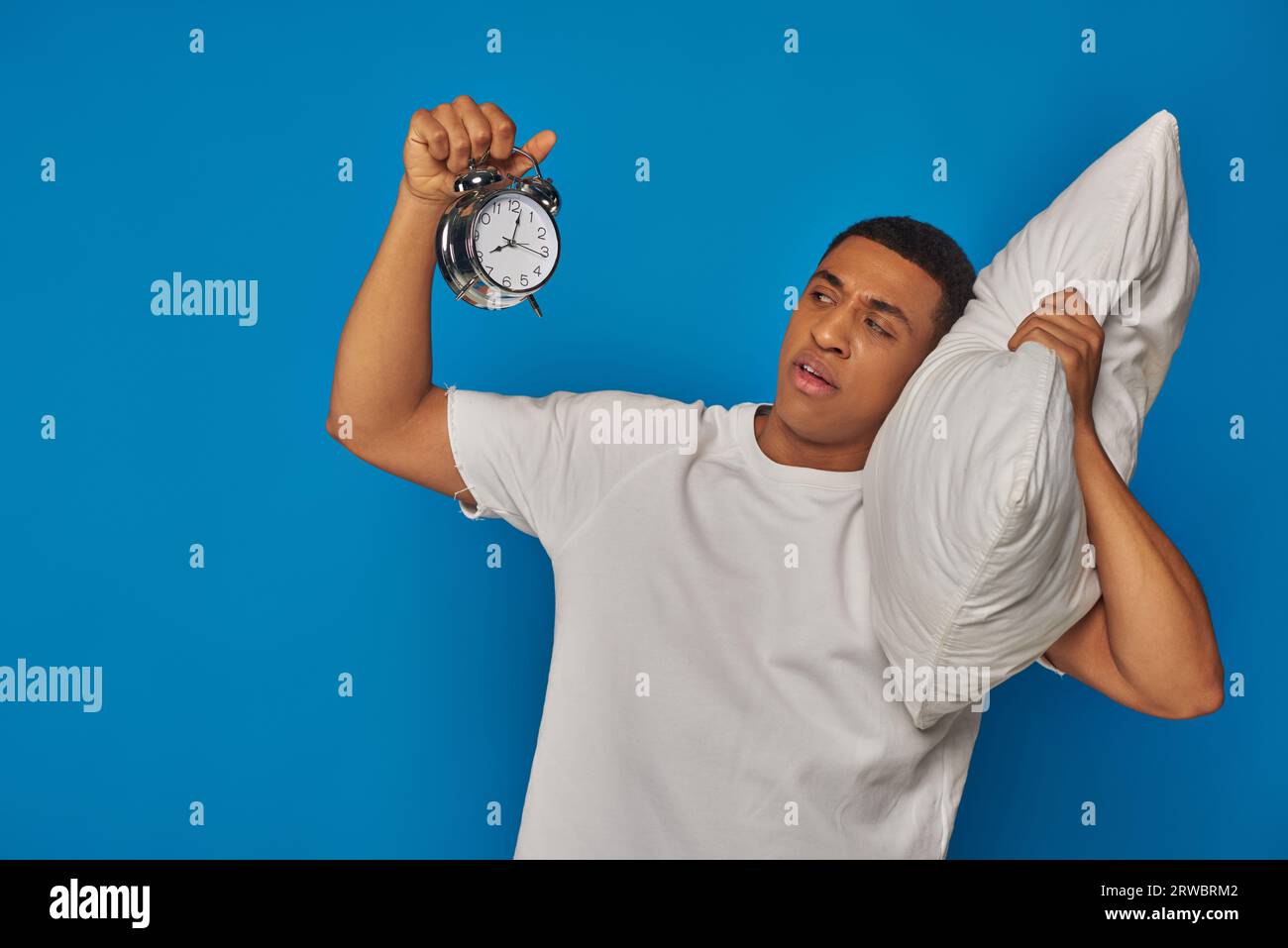 sleepy african american man lying on pillow and looking at retro alarm clock on blue background Stock Photo