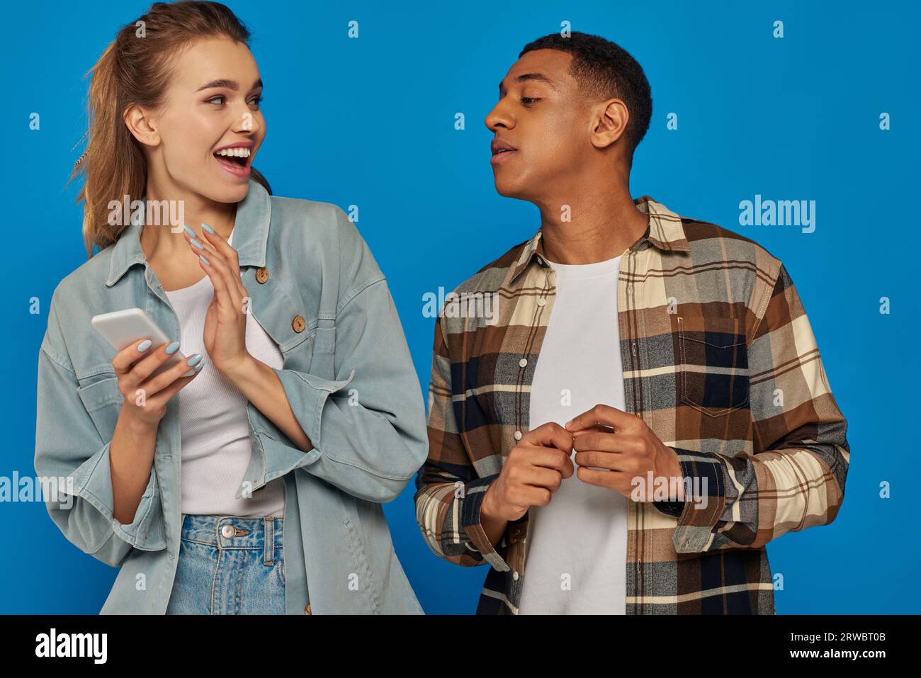 playful young woman hiding screen of smartphone from african american friend on blue backdrop Stock Photo