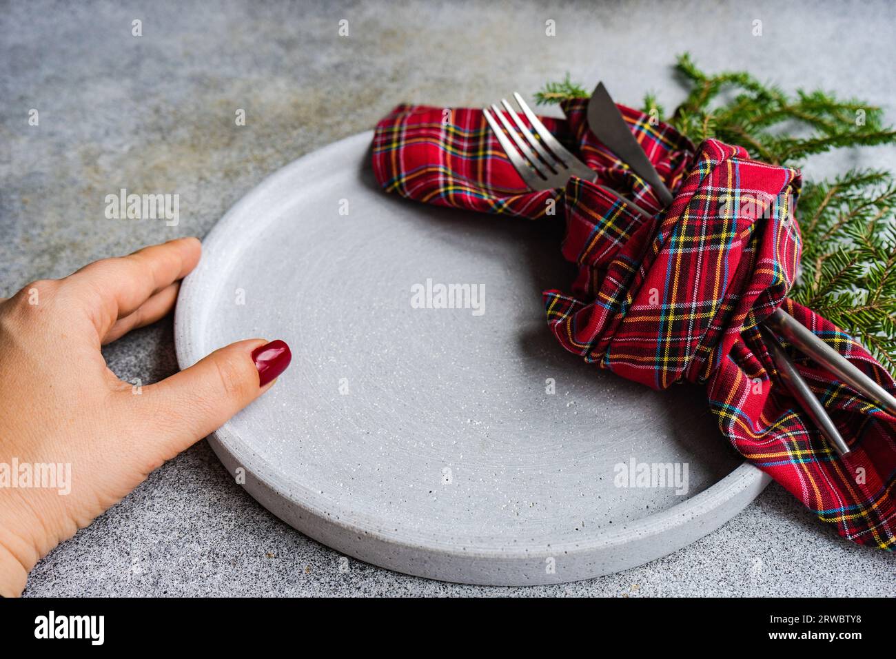 Unrecognizable female putting ceramic plate with fork and knife wrapped in checkered handkerchief on gray table near green fir sprigs Stock Photo