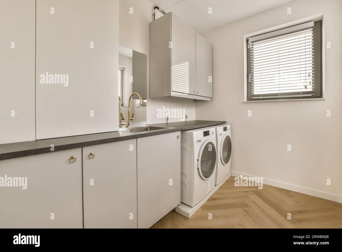 Empty white laundry room with modern washing and dryer machines placed by cabinets and sink in light modern kitchen Stock Photo