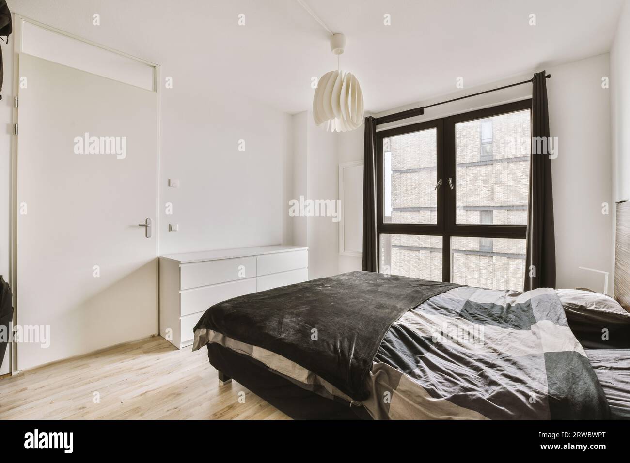 Light hanging on ceiling over comfortable bed next to large window with white walls and door in bedroom at contemporary apartment during daytime Stock Photo