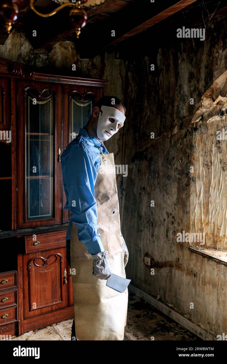 Anonymous terrifying man in butcher costume with apron and knife looking at camera while standing in old living room near window at home Stock Photo