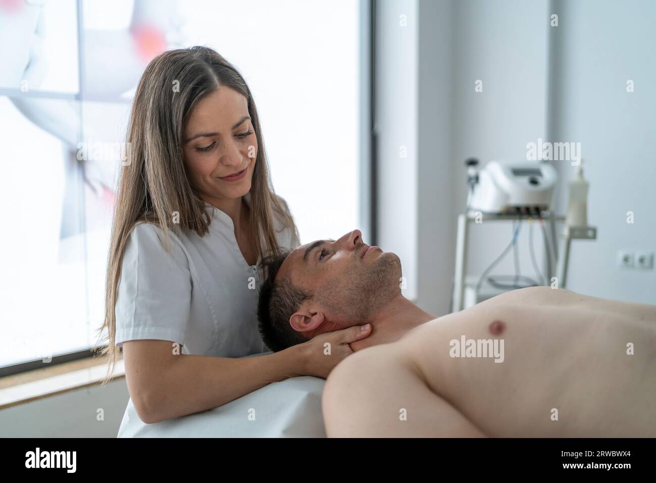 Positive young female therapist in white uniform smiling and massaging neck of calm male patient lying on table without shirt during physiotherapy ses Stock Photo
