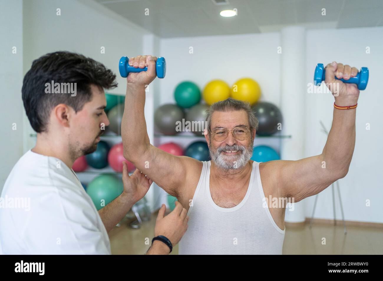 Serious male therapist in uniform helping elderly patient to do exercise with dumbbells during rehabilitation process in modern clinic center Stock Photo
