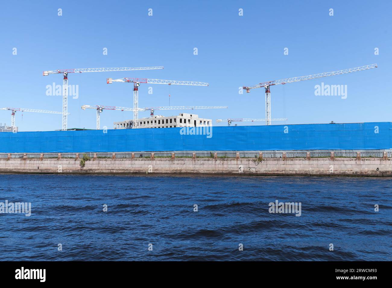 White tower cranes under clear blue sky, construction site is behind blue metal fence at the coast of Neva river, St-Petersburg, Russia Stock Photo
