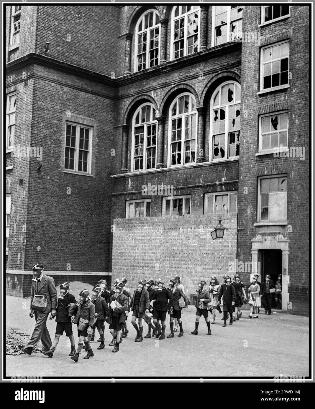 THE BLITZ LONDON WW2 Anti-gas drill at Old Woolwich Road School in Greenwich, London 1941. A school teacher wearing a gas mask leads group of school children out of the entrance of Old Woolwich Road School in Greenwich, London. The children are crossing playground, all are wearing their gas masks. The school has clearly been damaged by Nazi Germany blitz air raids, many windows have panes missing or smashed. 1941 London Blitz Battle of Britain World War II Stock Photo
