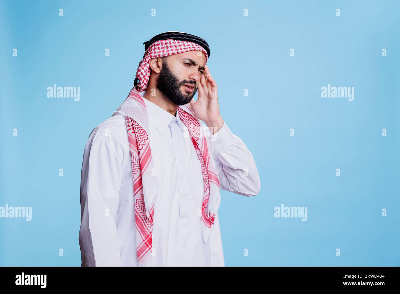 Muslim man wearing traditional clothes suffering from migraine and rubbing temple. Arab person in thobe and headscarf touching head while having headache and posing in studio Stock Photo