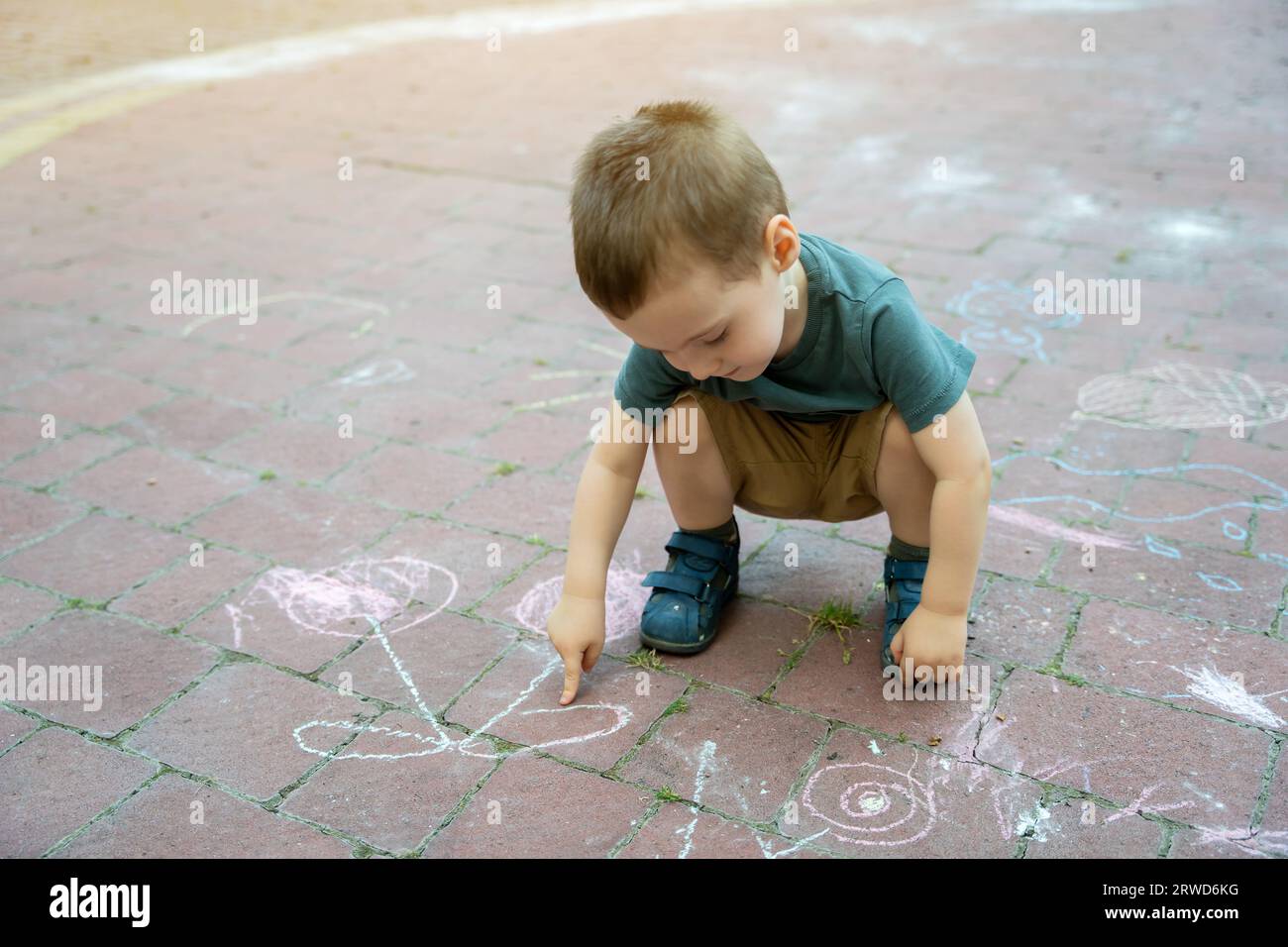 Little toddler boy two or three years old points his finger at children's drawings drawn in chalk. Playtime for children. Children's games. Selective Stock Photo