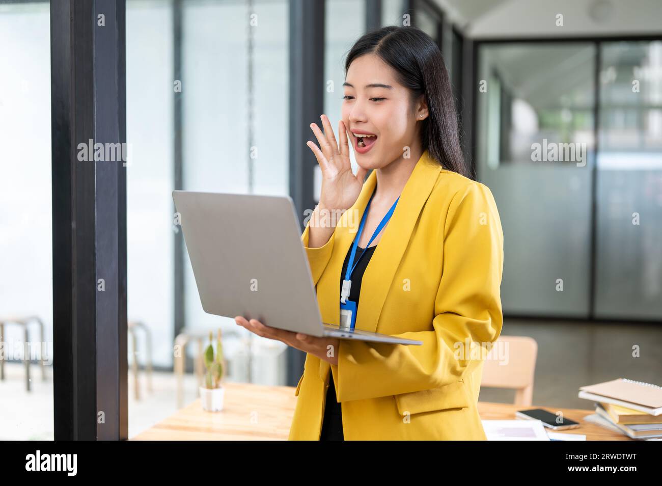 A cheerful and excited millennial Asian businesswoman in a yellow suit looking at her laptop screen with an excited face while standing in her office. Stock Photo