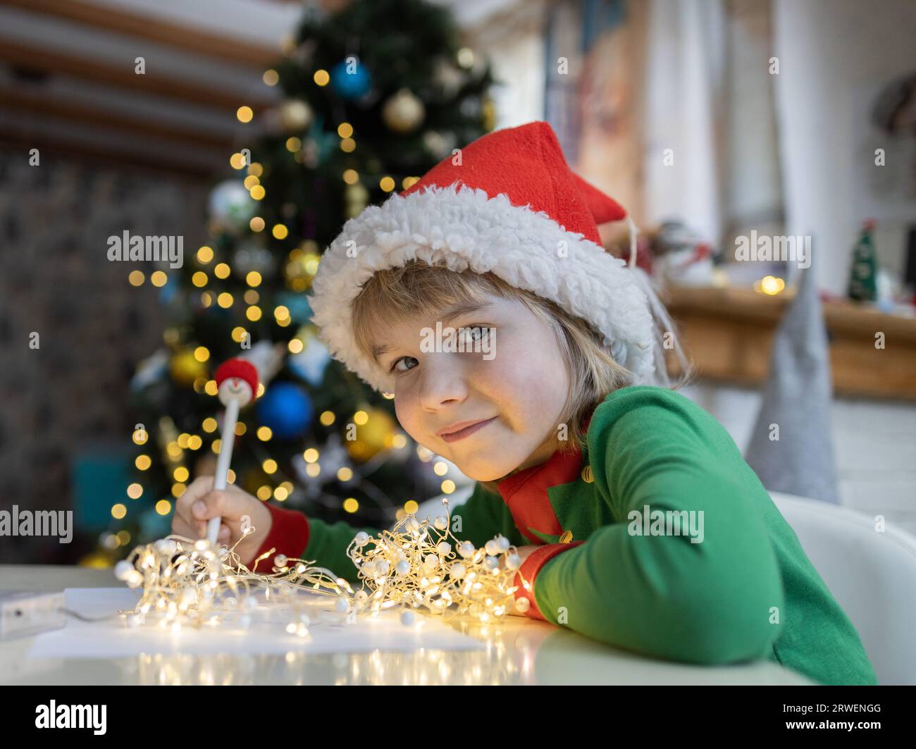 Christmas child writes letter to Santa while sitting at table against a bokeh background. 7 year old boy in red Santa hat dreams of gifts at home. Gif Stock Photo