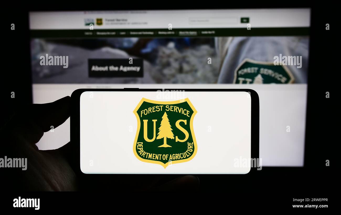 Person holding cellphone with logo of United States Forest Service (USFS) on screen in front of webpage. Focus on phone display. Stock Photo