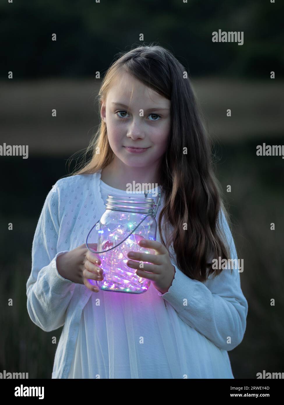 Close-up portrait of a young girl looking at the camera, holding a glass jar with fairy lights at dusk Stock Photo