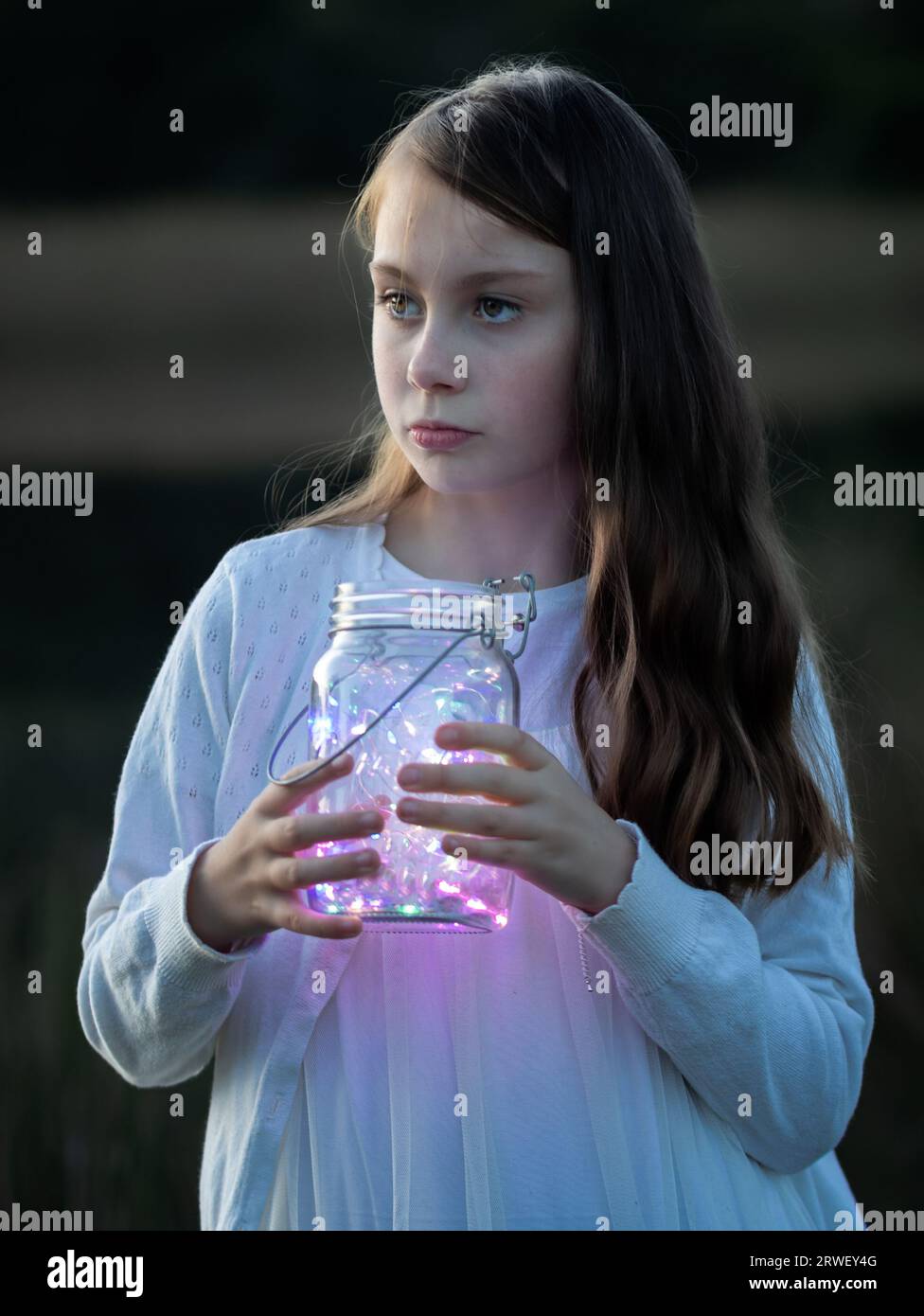 Close-up portrait of a young girl holding a glass jar with fairy lights at dusk looking to the side Stock Photo