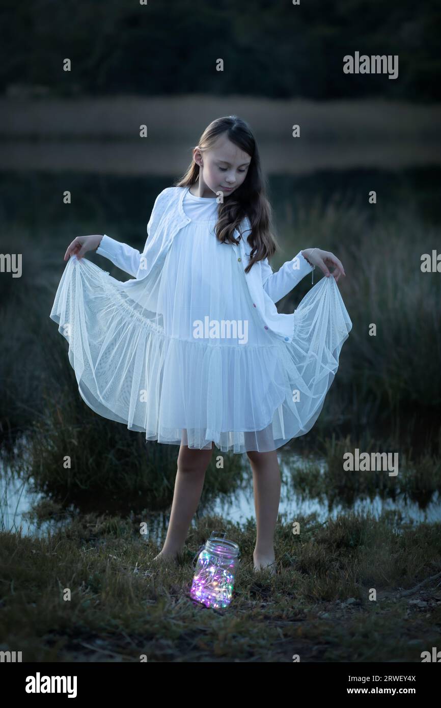 Side lit full length portrait of a young girl dressed in a white dress at dusk looking down at dusk Stock Photo