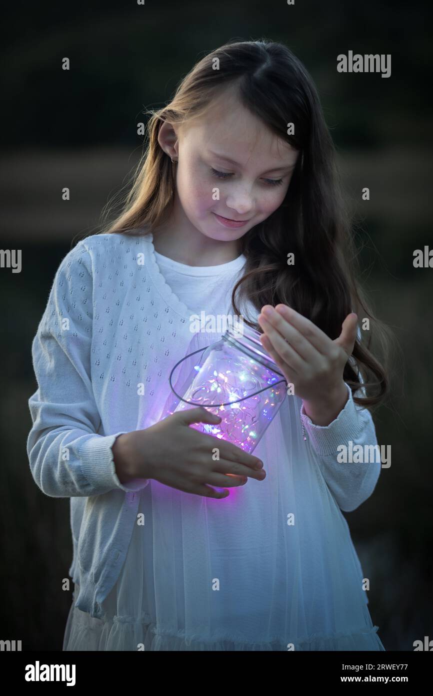 Close-up portrait of a young girl holding a glass jar with fairy lights at dusk looking down Stock Photo
