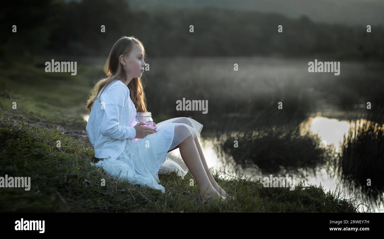 Portrait of a young girl holding a lantern sitting at the water's edge covered in a soft mist at dusk Stock Photo