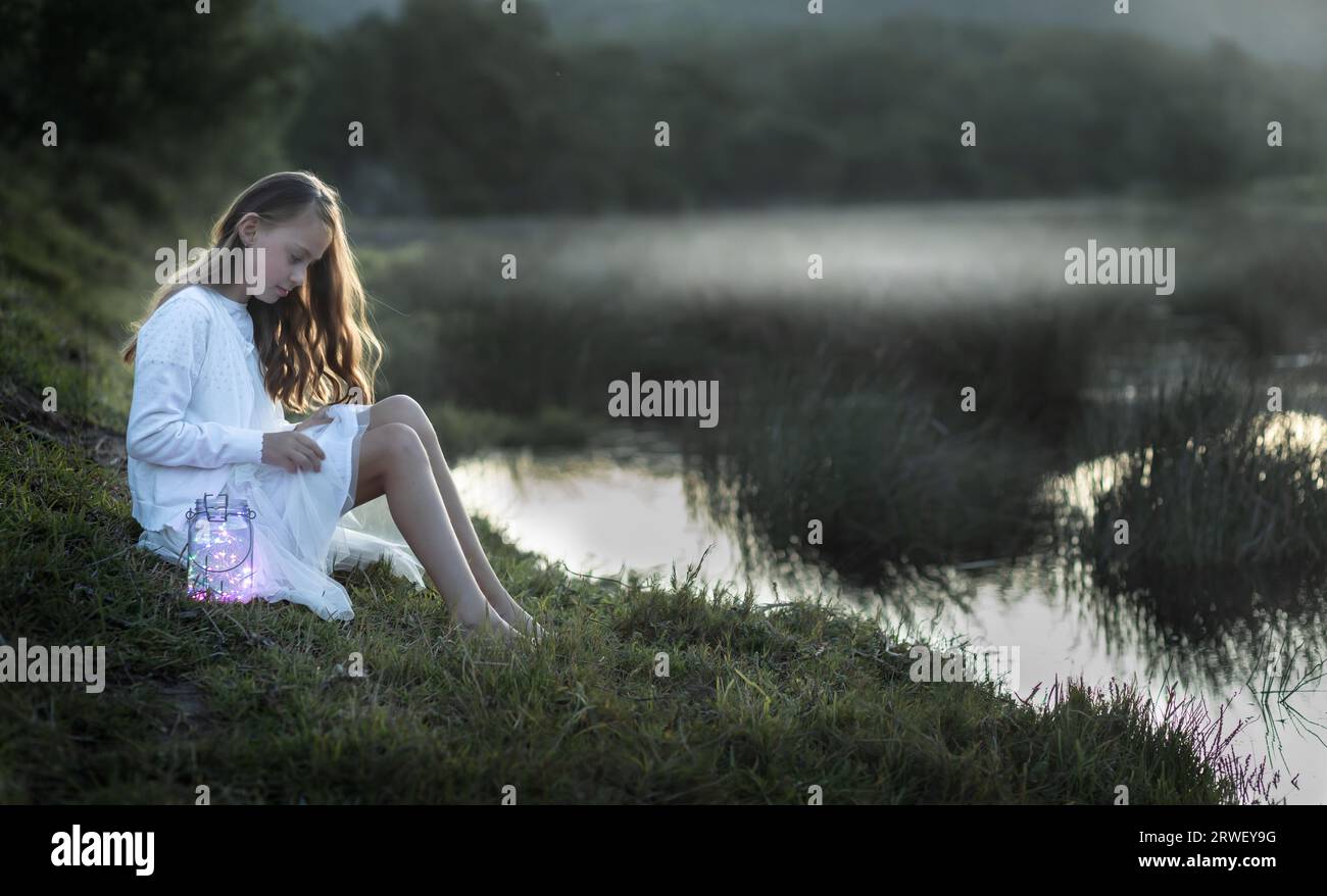 Back lit portrait of a young girl dressed in white sitting at the water's edge looking down Stock Photo
