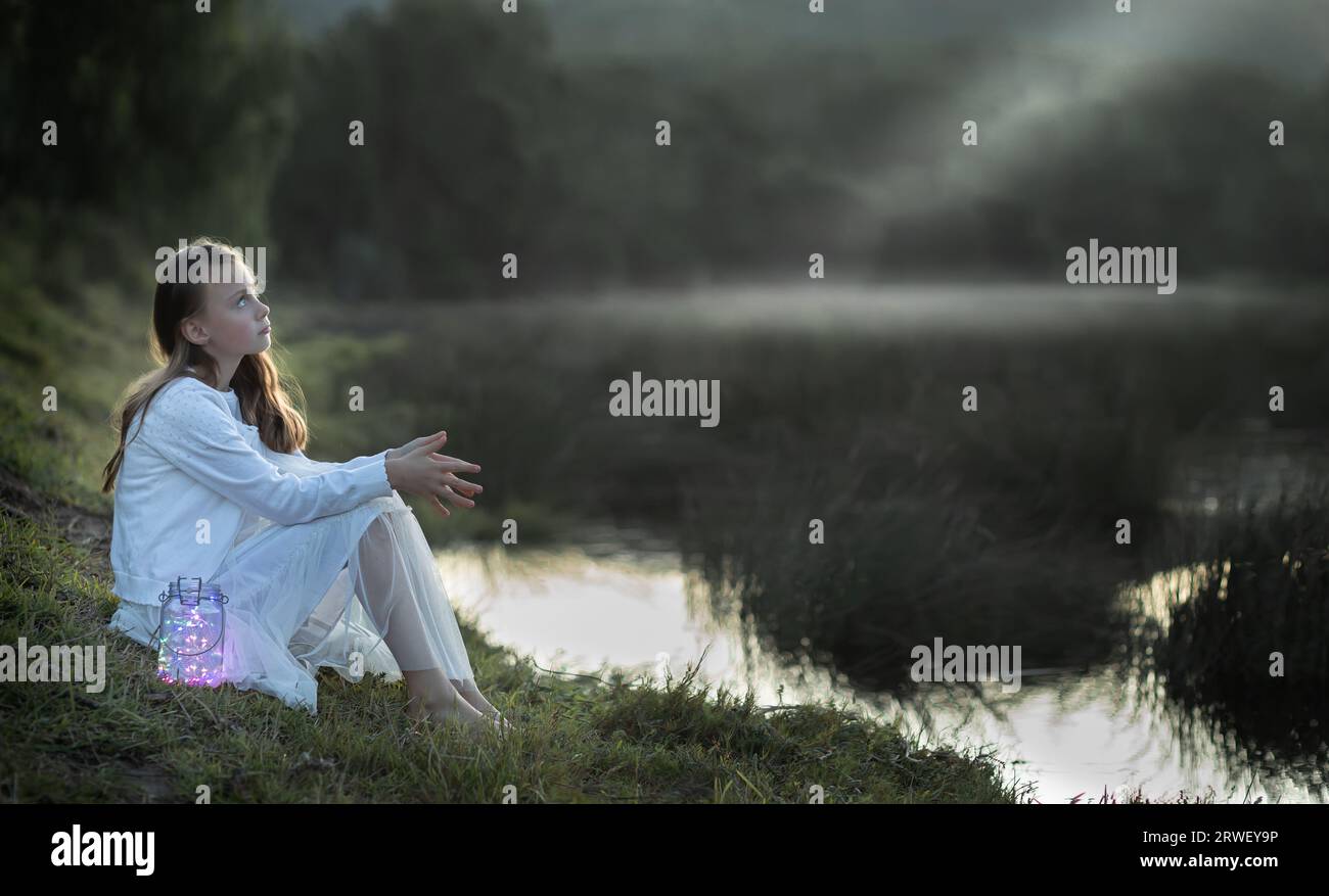 Portrait of a young girl dressed in white sitting at the water's edge looking up at dusk Stock Photo
