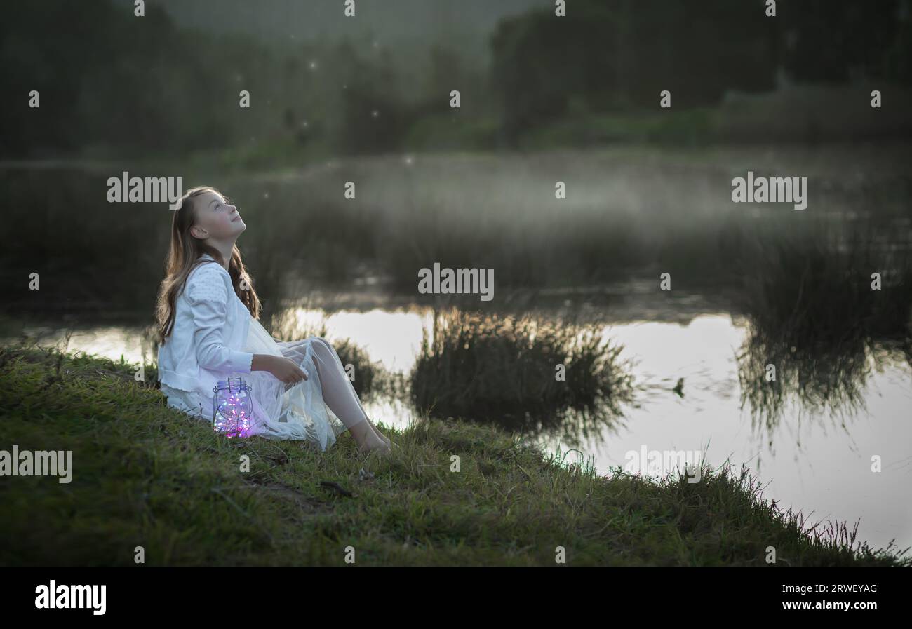 Portrait of a young girl dressed in white sitting at the water's edge looking up at fireflies Stock Photo