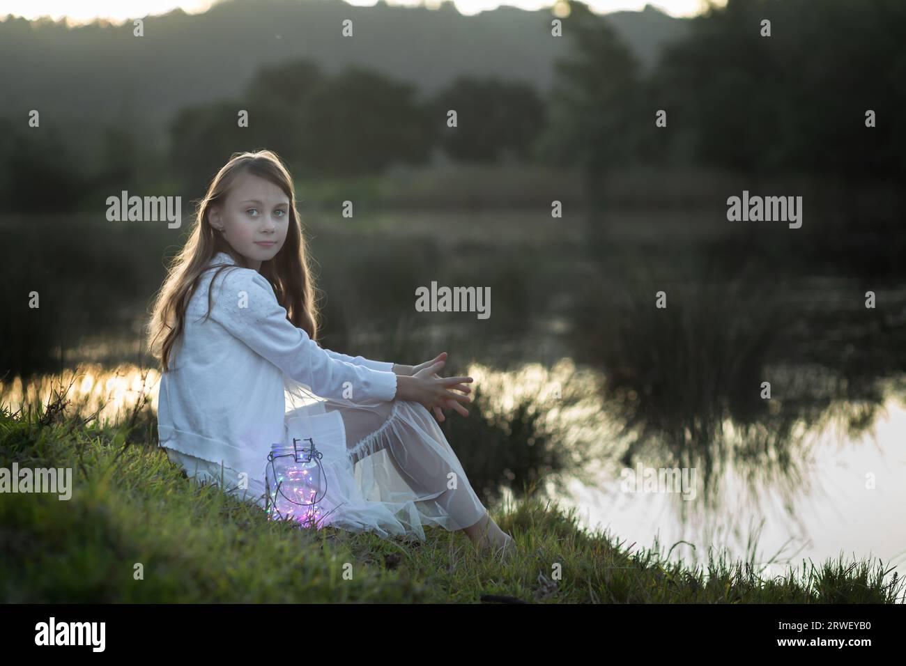 Back lit portrait of a young girl dressed in white sitting at the water's edge looking at the camera Stock Photo