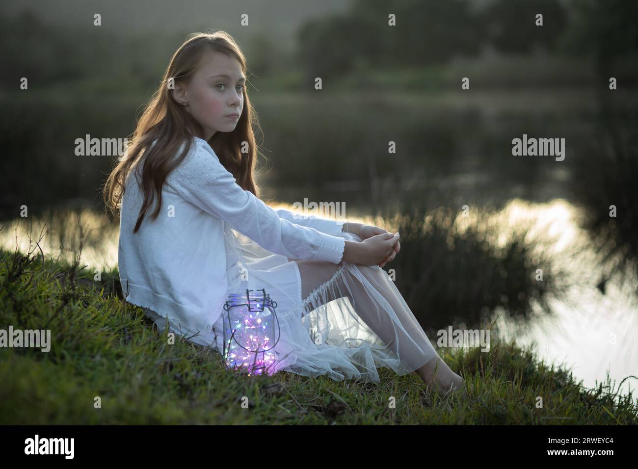 Back lit portrait of a young girl dressed in white sitting at the water's edge looking at the camera Stock Photo