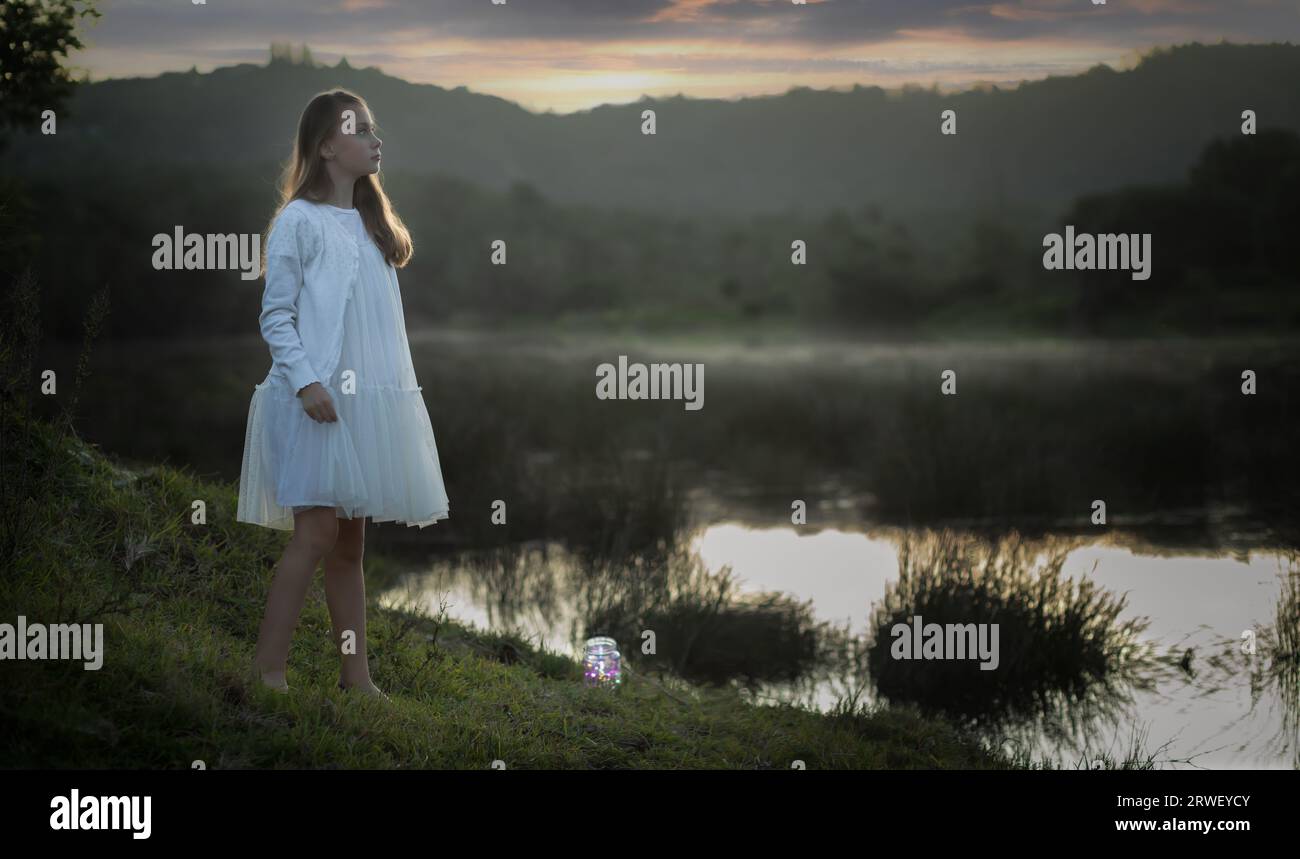 Portrait of a young girl in a white dress standing at the edge of a lake at sunset Stock Photo