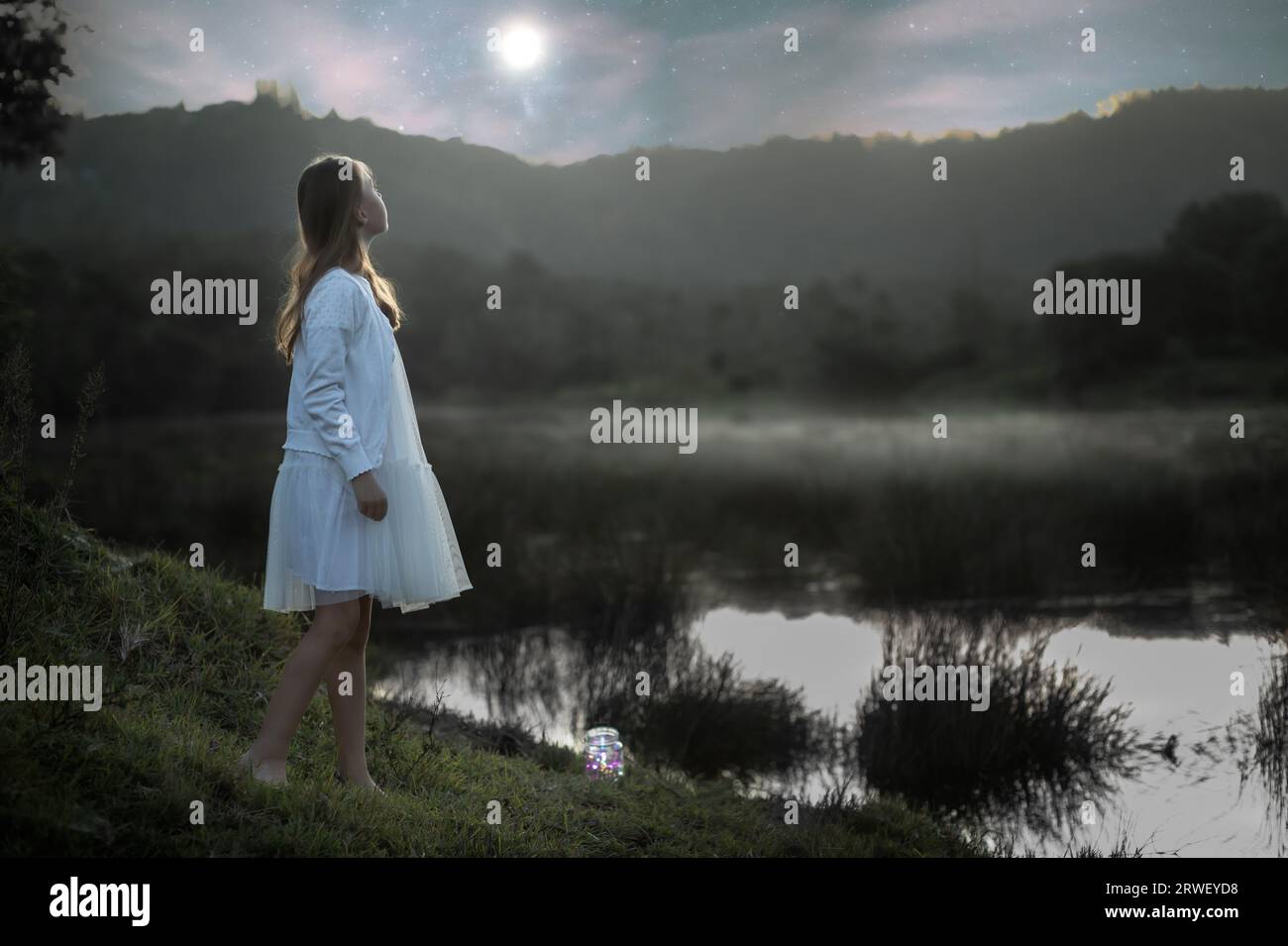 Back lit portrait of a young girl dressed in white standing at the water's edge at dusk looking at the moon rising over the mountain Stock Photo