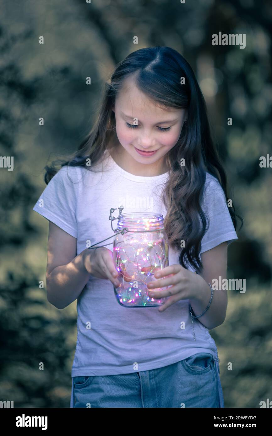 Young girl with long hair, dressed in a white top, holding a lantern of fairy lights at dusk Stock Photo