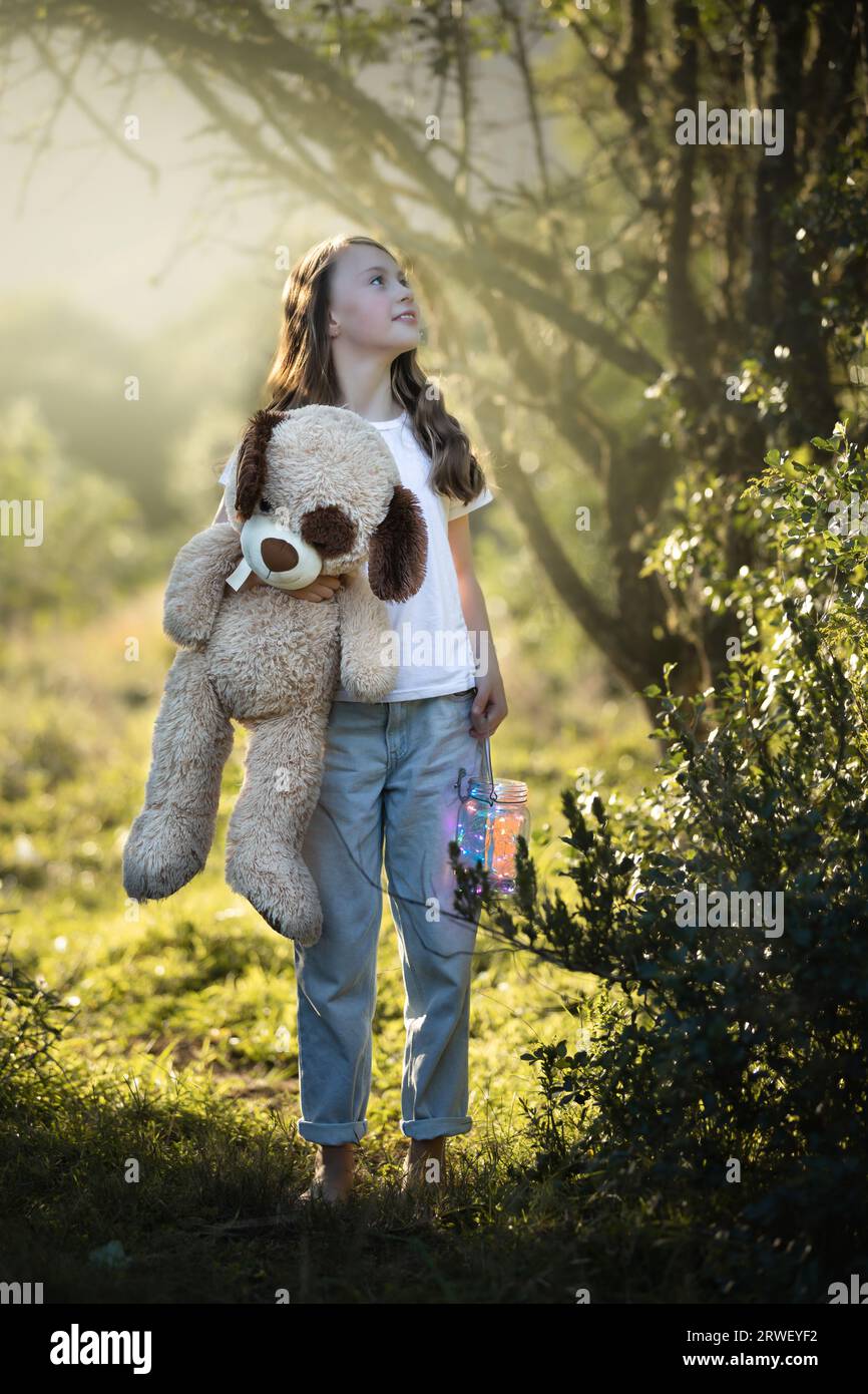 Young girl standing under trees, holding her teddy and lantern at sunset Stock Photo