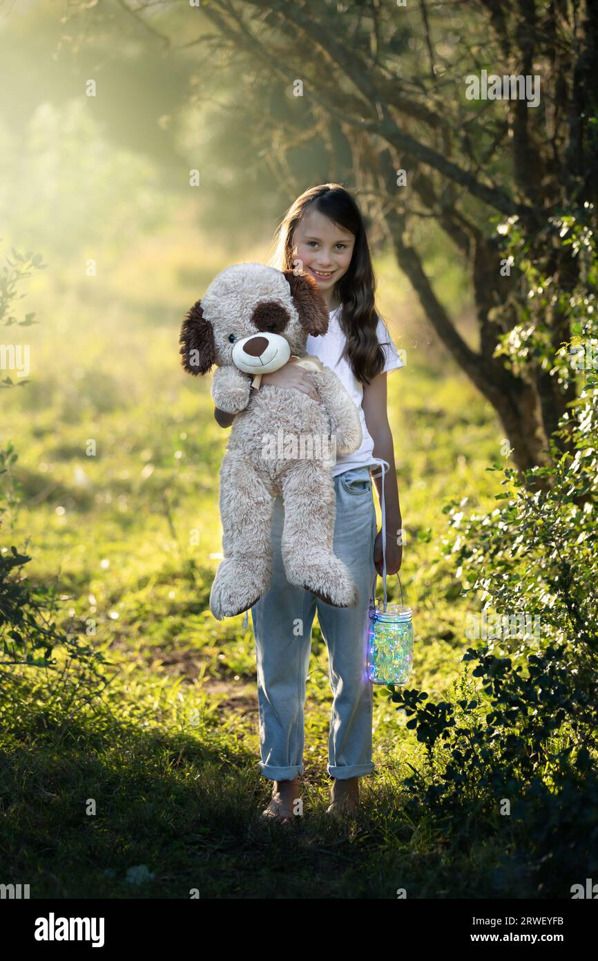 Young girl standing under trees, holding her teddy and lantern at sunset Stock Photo