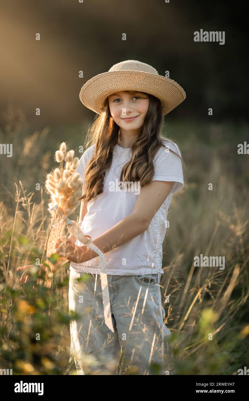 Young girl casually dressed and wearing a hat is standing in a field holding a posy of dried grass Stock Photo