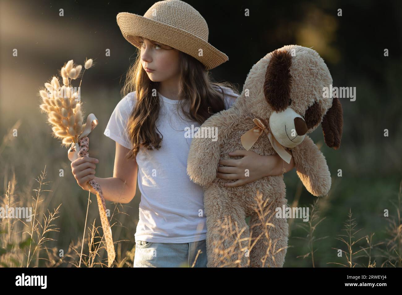 Young Caucasian girl wearing a hat and holding a posy of dried grass and her teddy bear whilst looking sideways at sunset Stock Photo