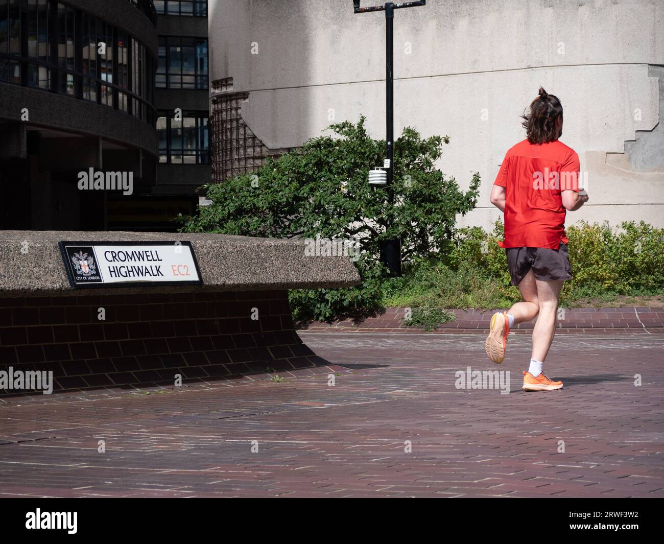 Runner passing Cromwell Highwalk sign, The Barbican Estate, or Barbican, residential area of around 2,000 flats, maisonettes, and houses in City of London , England, with crumbling wall in background Stock Photo