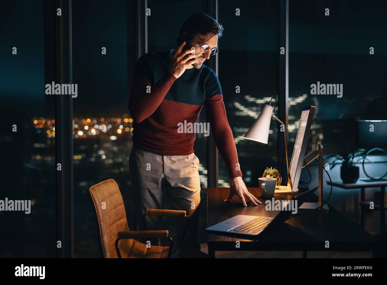 businessman standing at his desk in an office, talking on his mobile phone as he works late into the night. Professional man communicating with his co Stock Photo