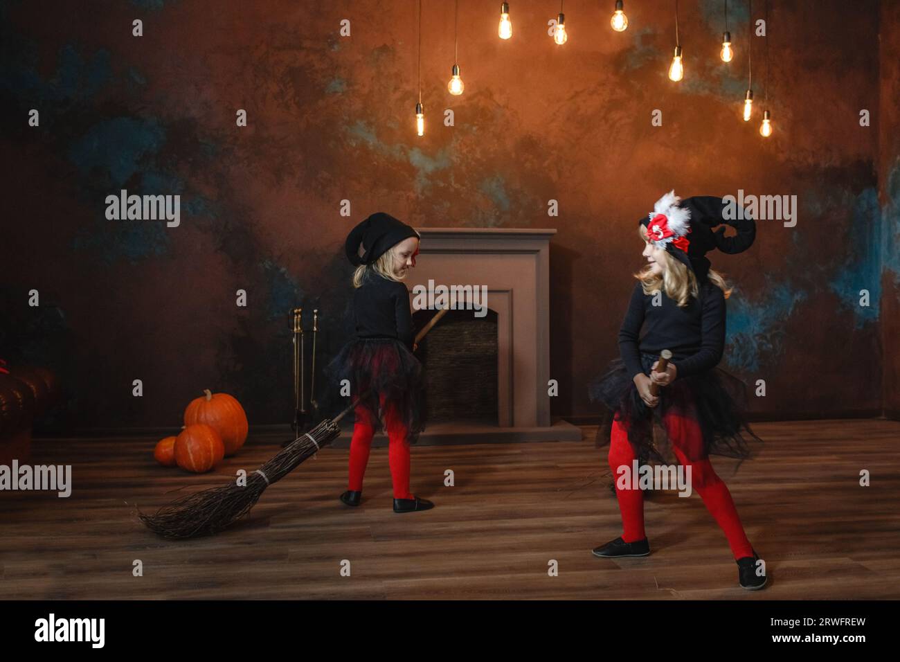 Little witches sit on brooms, run around the room one after another, and play. Lanterns glow in the background. Halloween. Stock Photo