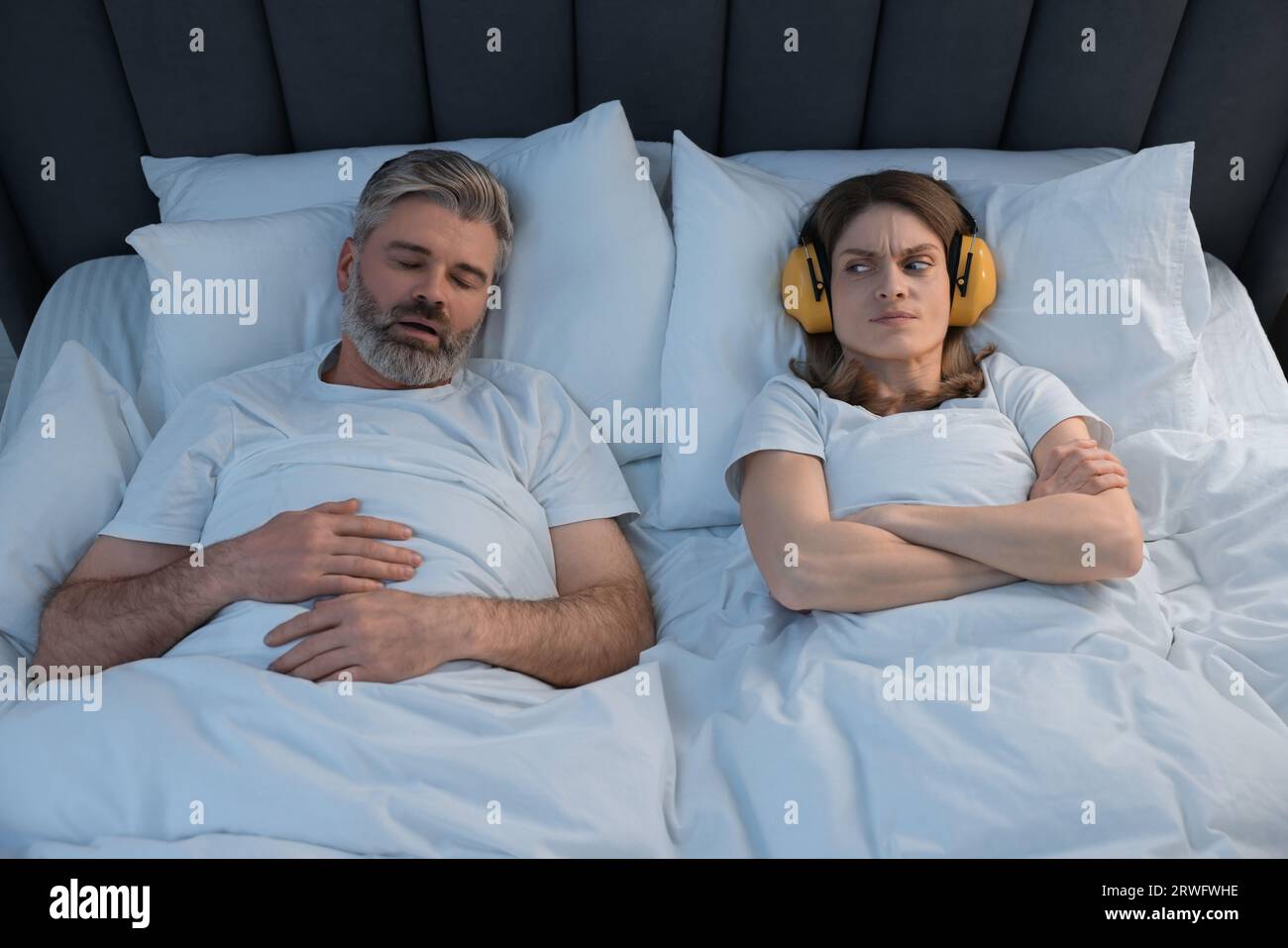 Irritated woman with headphones lying near her snoring husband in bed at home Stock Photo