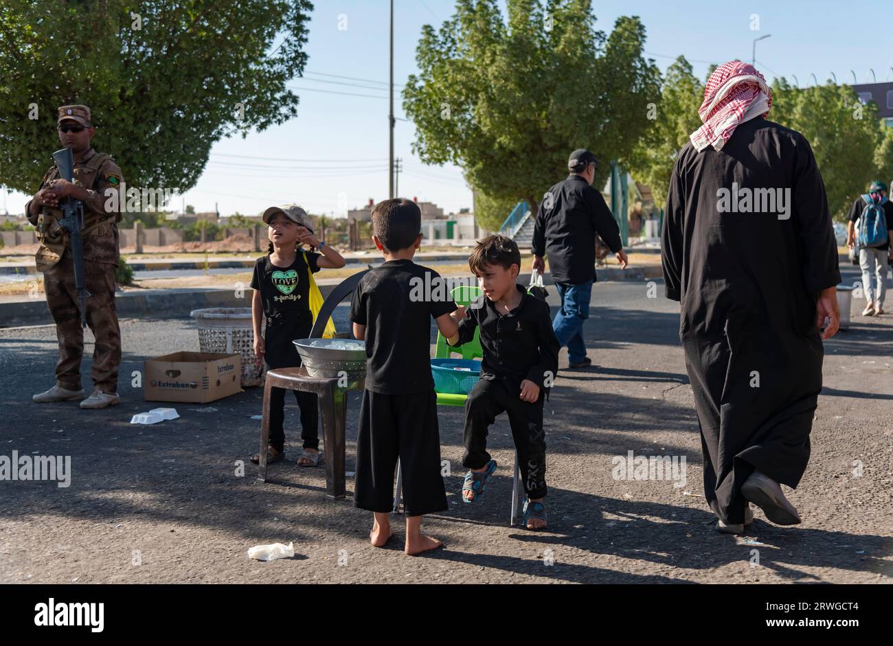 Iraqi Shia kids distribute water among Shia Muslim pilgrims marching from Najaf towards Shrine city of Karbala. Every year, millions of Shia Muslims and some from other faiths undertake a 20-day pilgrimage on foot from various cities in Iraq and Iran to the holy city of Karbala. This pilgrimage is in remembrance of Imam Hussein, the grandson of the Prophet Muhammad, who died in a battle in 680 AD. On the 40th day of mourning for Hussein, known as Arbaeen, pilgrims converge in Karbala to pay tribute at his shrine. Along the way, volunteers provide food, water, and shelter, and villagers open th Stock Photo