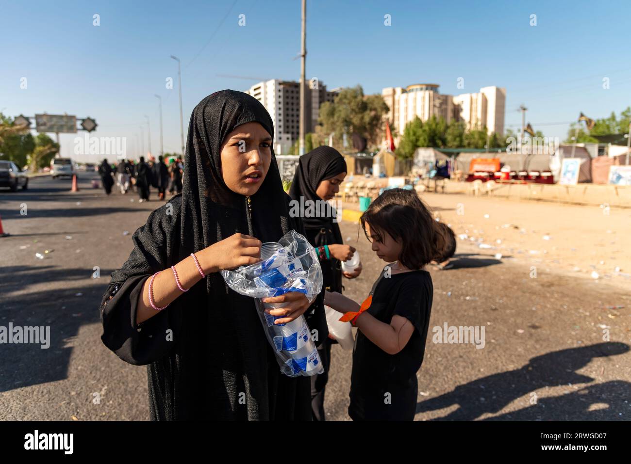 An Iraqi Shia girl distribute juice among Shia Muslim pilgrims marching from Najaf towards Shrine city of Karbala. Every year, millions of Shia Muslims and some from other faiths undertake a 20-day pilgrimage on foot from various cities in Iraq and Iran to the holy city of Karbala. This pilgrimage is in remembrance of Imam Hussein, the grandson of the Prophet Muhammad, who died in a battle in 680 AD. On the 40th day of mourning for Hussein, known as Arbaeen, pilgrims converge in Karbala to pay tribute at his shrine. Along the way, volunteers provide food, water, and shelter, and villagers open Stock Photo