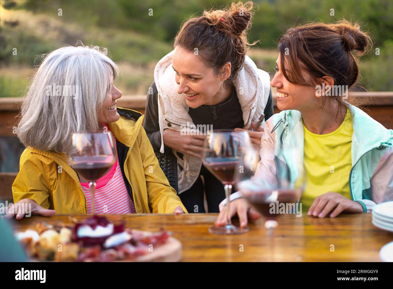 Three middle-aged friends chat on a mountain terrace at sunset with wine glasses and a cutting board nearby. One with gray hair, two with tied hair, a Stock Photo